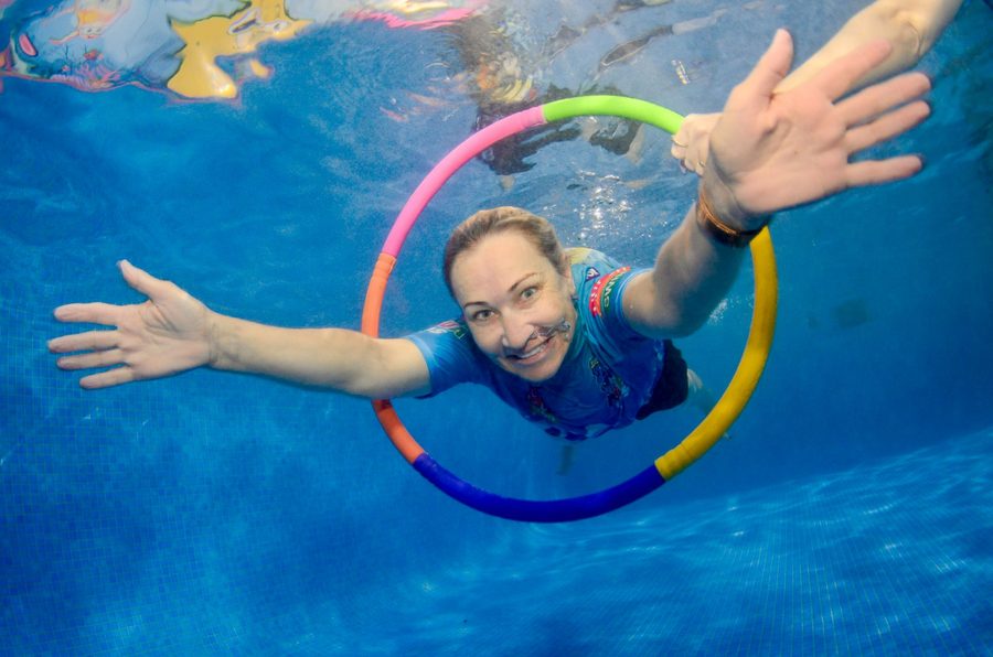Angela Rothwell underwater in the AQUAKids pool, arms outstretched and smiling at the camera as she swims through a coloured hoop.
