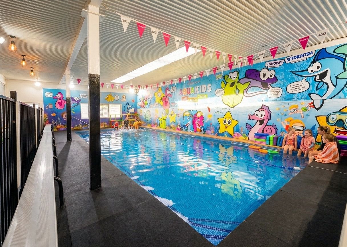 The indoor pool at AQUAKids Smithfield, with the mascot mural along the far wall and children waiting on the deck in towels.