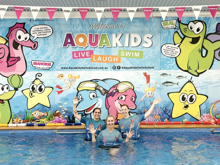 Three AQUAKids instructors in team shirts waving from the pool, in front of the painted mascot mural.